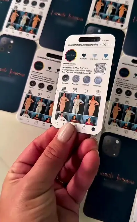 Cartão de visita no formato de celular, com o visual de um perfil do Instagram, na mão, sobre uma pilha de cartões